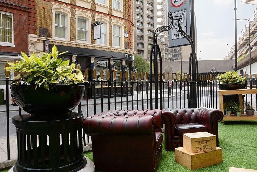 فندق Montcalm Brewery Townhouse لندن المظهر الخارجي الصورة The photo shows a street scene with a cozy outdoor seating area. There are two plush, dark leather couches arranged on green grass, creating a welcoming atmosphere. Surrounding the seating are large planters with vibrant plants. In the background, th