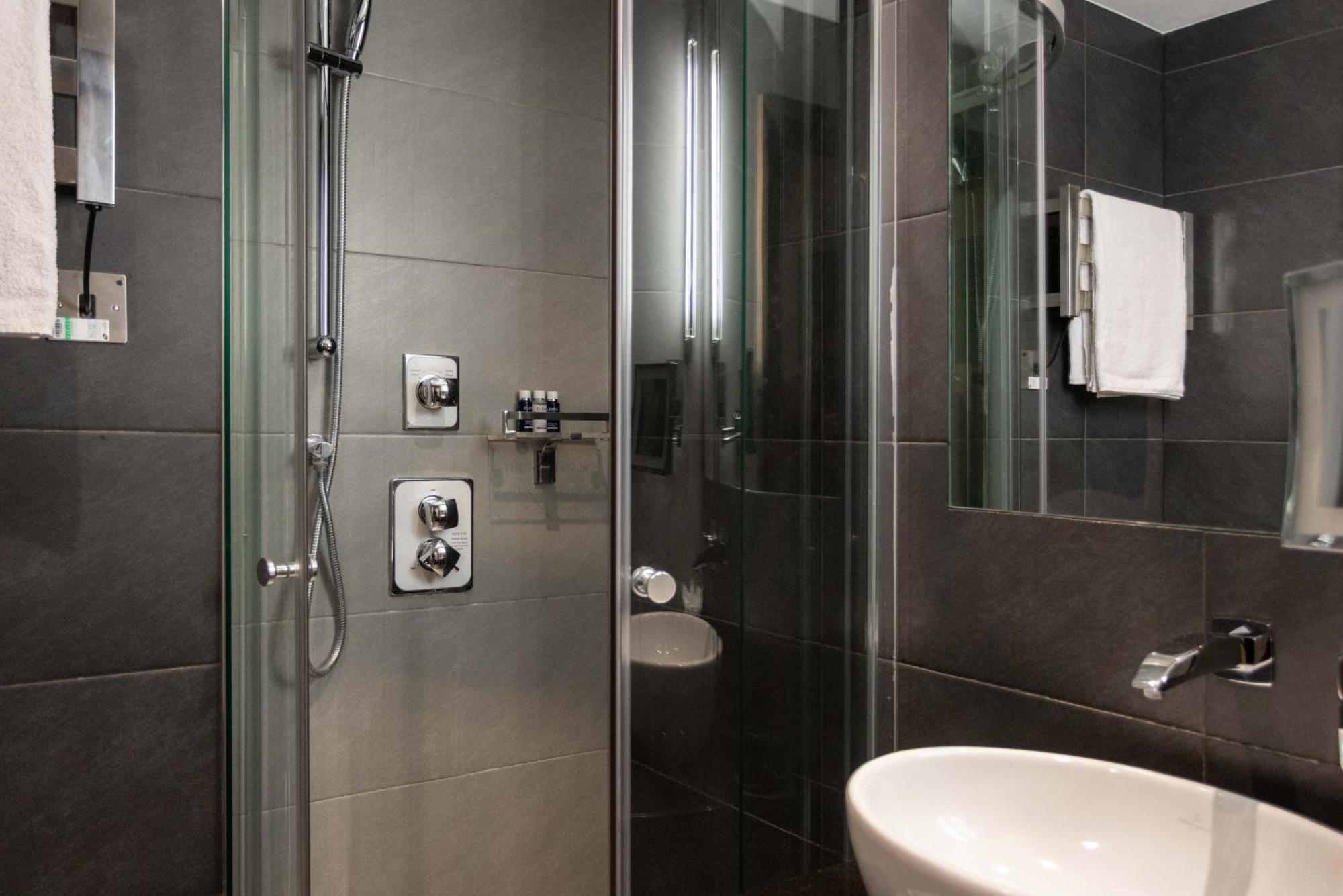 فندق Montcalm Brewery Townhouse لندن المظهر الخارجي الصورة The photo shows a modern bathroom with a glass shower enclosure. The shower features a handheld showerhead and various controls mounted on the wall. The walls are tiled in dark, sleek tones, contributing to a contemporary aesthetic. There is a round 