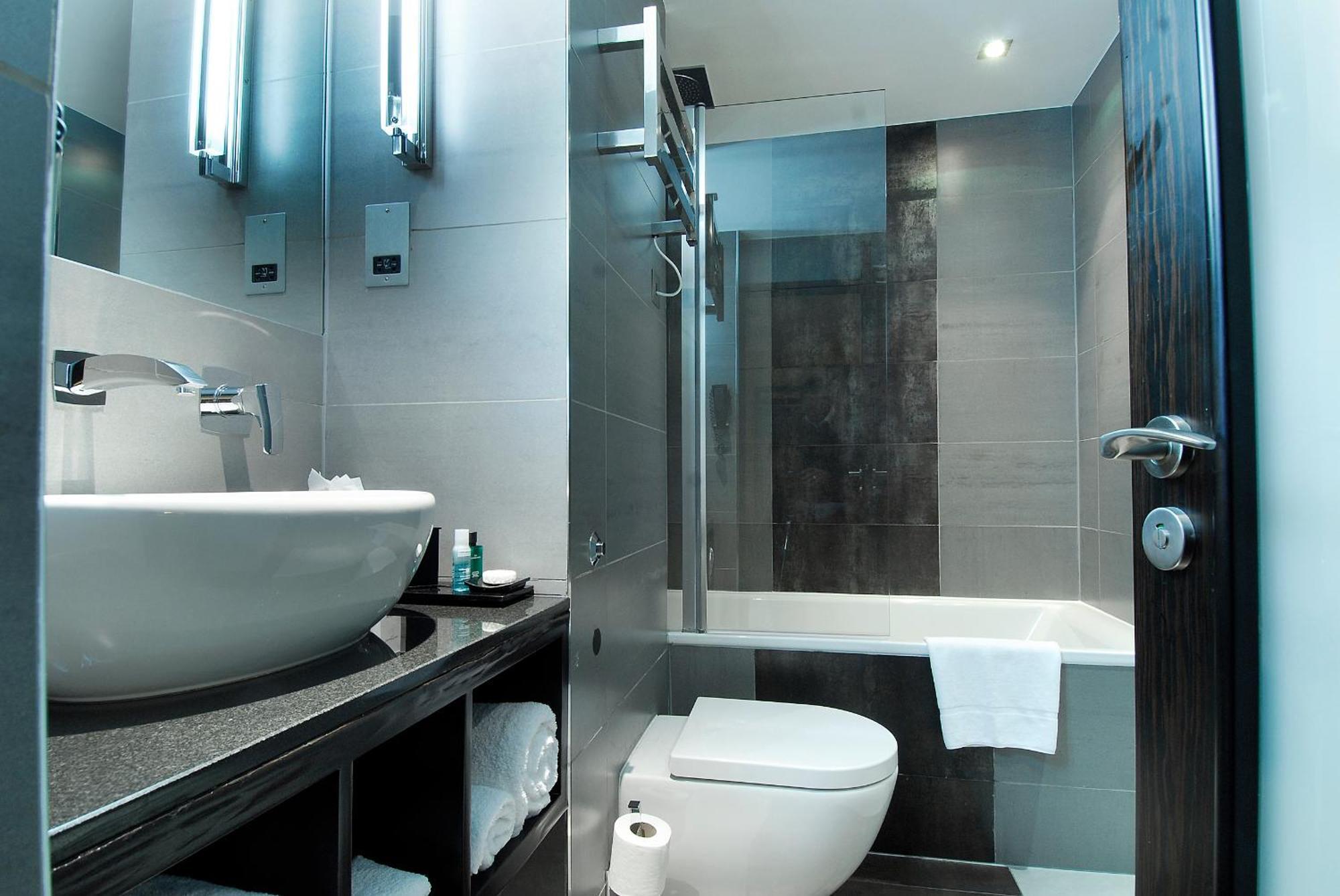 فندق Montcalm Brewery Townhouse لندن المظهر الخارجي الصورة The photo shows a modern bathroom with sleek design elements. It features a curved white sink on a dark countertop, with a mirror above that is illuminated by stylish lighting fixtures. Next to the sink, there's a toilet, and on the opposite side, a 