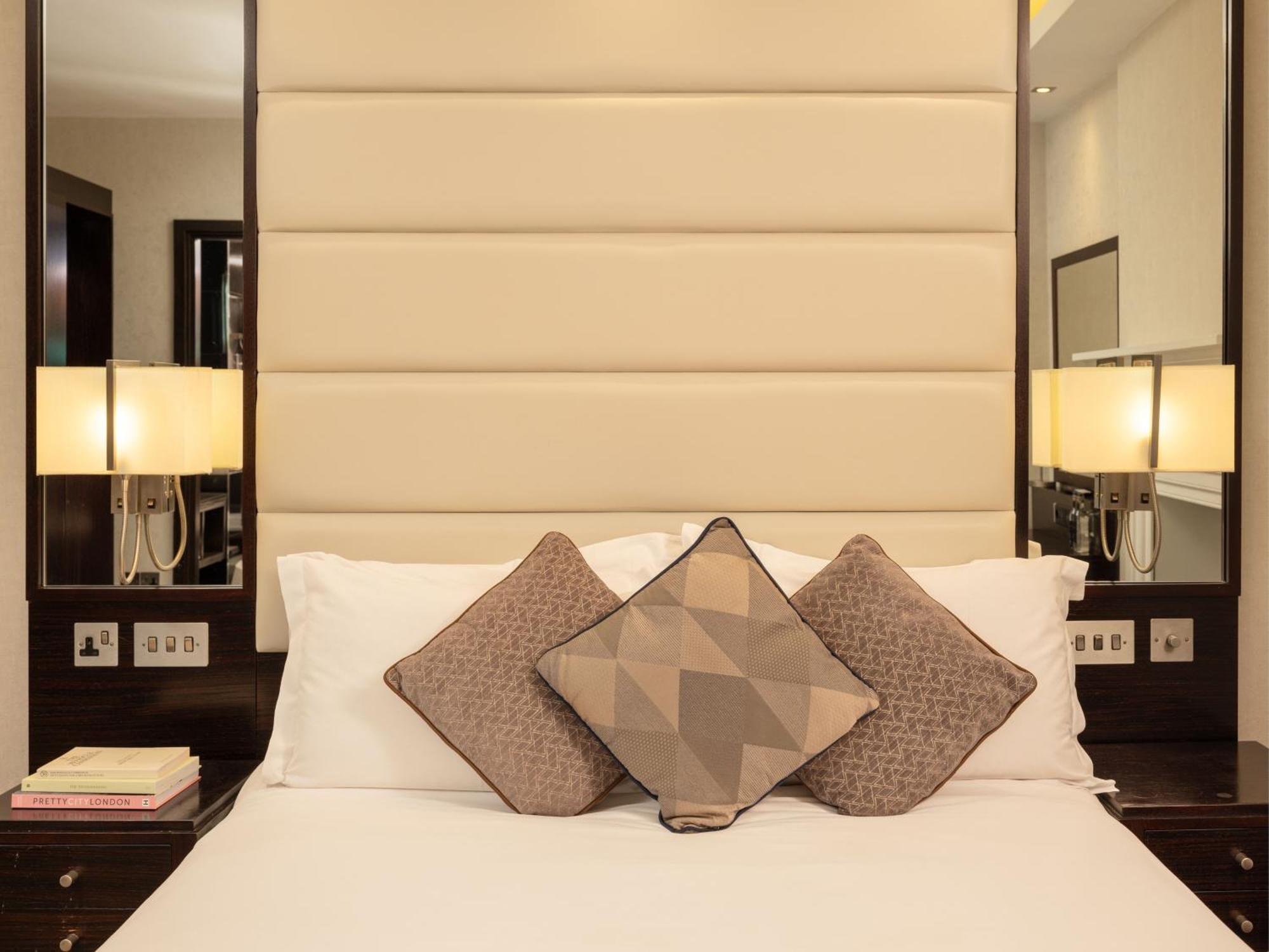 فندق Montcalm Brewery Townhouse لندن المظهر الخارجي الصورة The image shows a cozy bedroom setting. The bed features a headboard upholstered in a light beige fabric. It is adorned with three decorative pillows in varying patterns and colors, placed on white bedding. On either side of the bed, there are modern