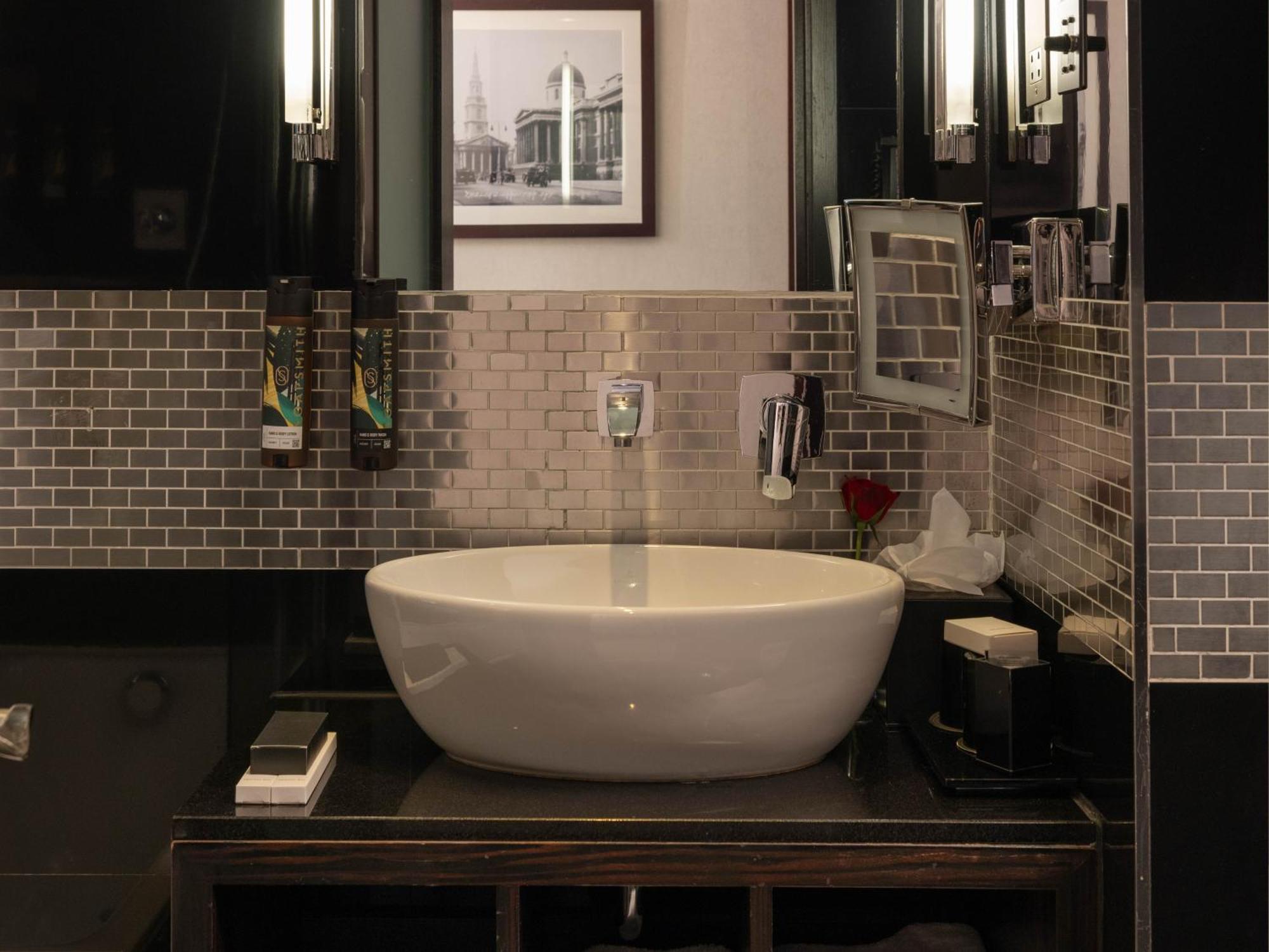 فندق Montcalm Brewery Townhouse لندن المظهر الخارجي الصورة The photo shows a modern bathroom with a stylish design. It features a large, round white sink placed on a dark wooden vanity. The walls are adorned with a mix of gray and silver tile, creating a sleek and contemporary look. Above the sink, there is 