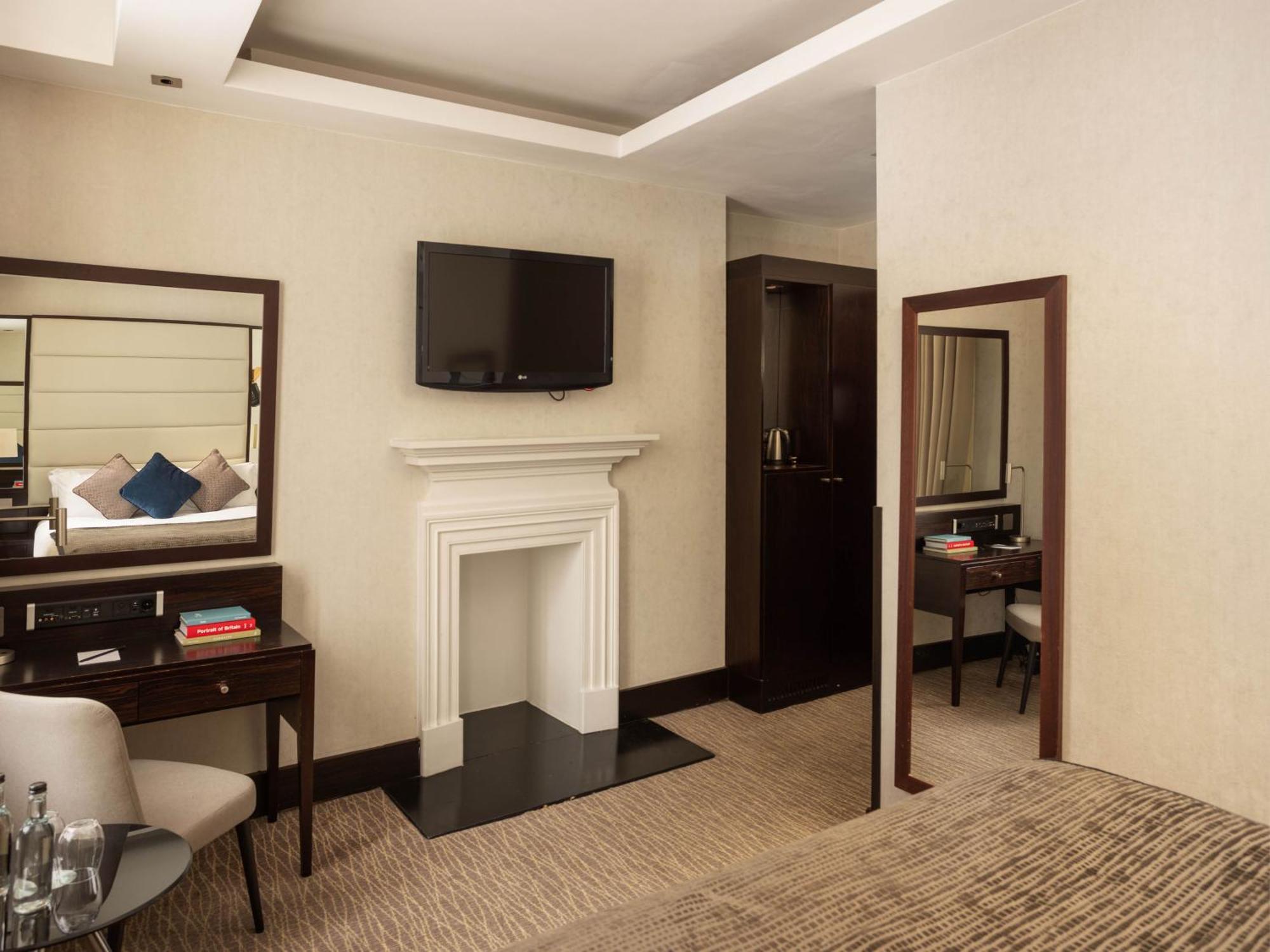 فندق Montcalm Brewery Townhouse لندن المظهر الخارجي الصورة The photo depicts a stylish hotel room. It features a double bed, a large flat-screen TV mounted on the wall, and a decorative fireplace. There is a wooden desk with a chair beside it, as well as a mirror hanging on the wall. The decor is modern and 