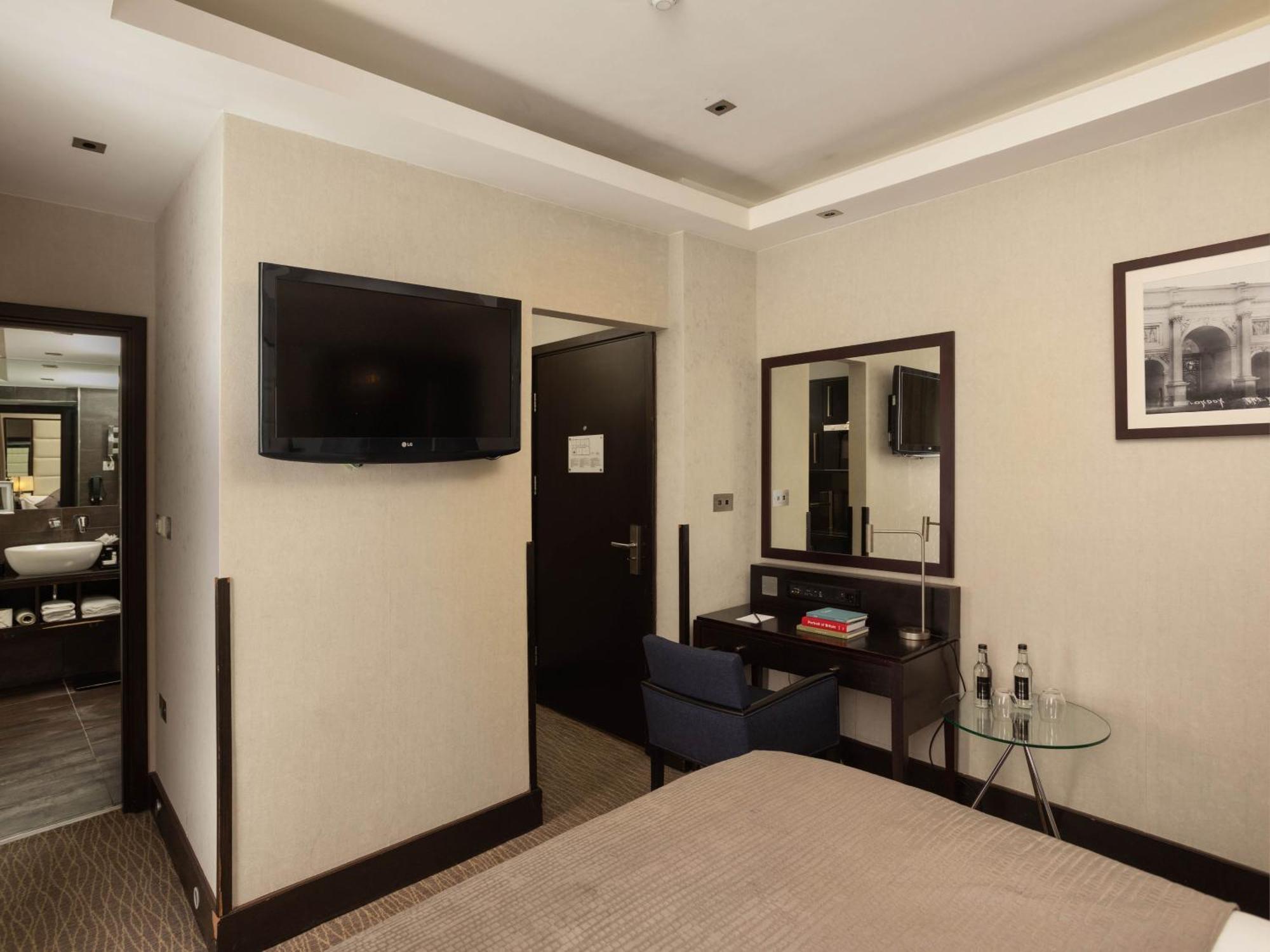 فندق Montcalm Brewery Townhouse لندن المظهر الخارجي الصورة The photo shows a hotel room with a modern and minimalist design. On the left, there's a wall-mounted flat-screen TV. In the center, there's a bed with a neutral-colored bedspread. To the right of the bed, there is a small desk with a mirror above it