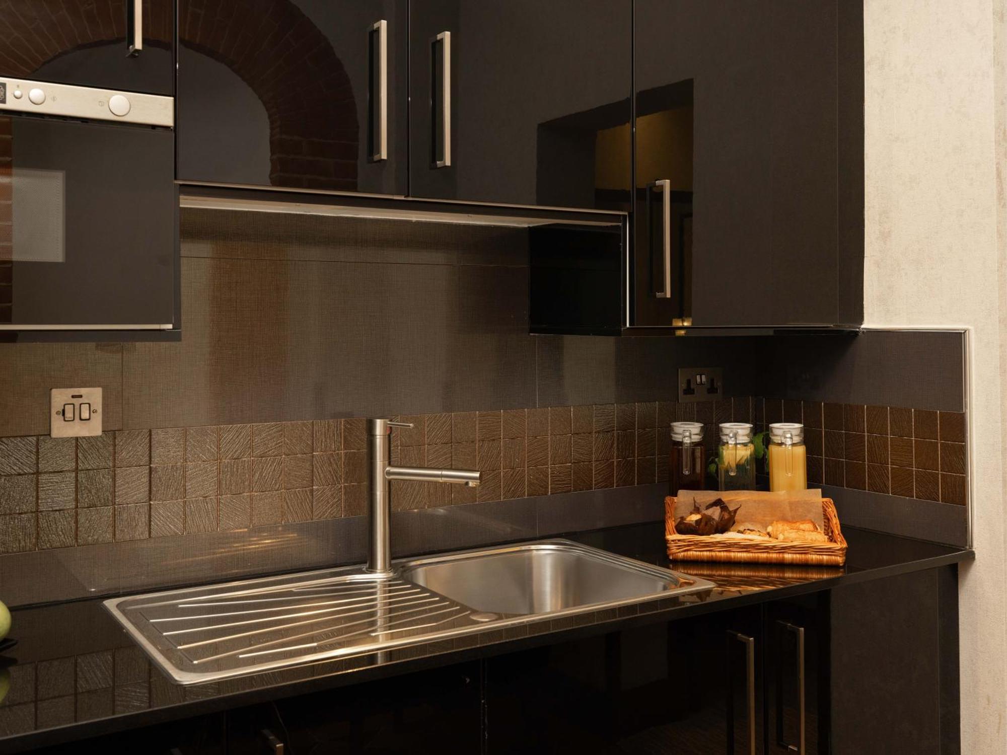 فندق Montcalm Brewery Townhouse لندن المظهر الخارجي الصورة The photo shows a modern kitchen with a sleek design. It features dark cabinetry with a glossy finish and stainless steel handles. There is a stainless steel sink with a simple faucet, and a countertop made of black material. Above the sink, there ar