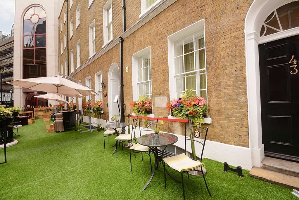فندق Montcalm Brewery Townhouse لندن المظهر الخارجي الصورة The photo depicts a charming outdoor area in front of a building with a warm, textured brick facade. There is a neatly maintained lawn, likely made of artificial grass, and several small round tables with chairs are arranged outside. Flower pots with