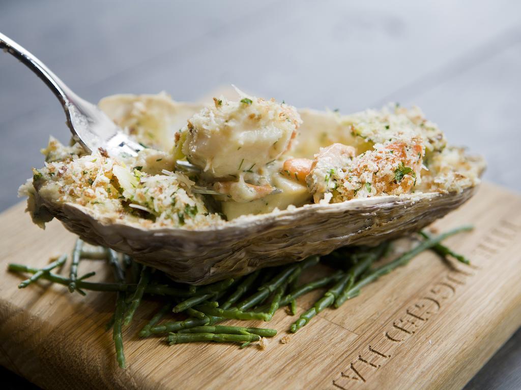 فندق Montcalm Brewery Townhouse لندن المظهر الخارجي الصورة The photo shows a large oyster shell filled with a mixture that appears to include seafood, herbs, and breadcrumbs. The dish is presented on a wooden board with some greenery, possibly a type of seaweed or garnishing. A fork is placed beside the oyst