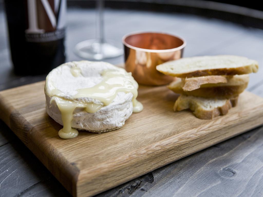 فندق Montcalm Brewery Townhouse لندن المظهر الخارجي الصورة The photo features a wooden serving board with several items. There is a round cheese, likely Brie or a similar soft cheese, that appears to be melting and dripping slightly. Next to the cheese, there is a small copper cup that might contain a sauce 