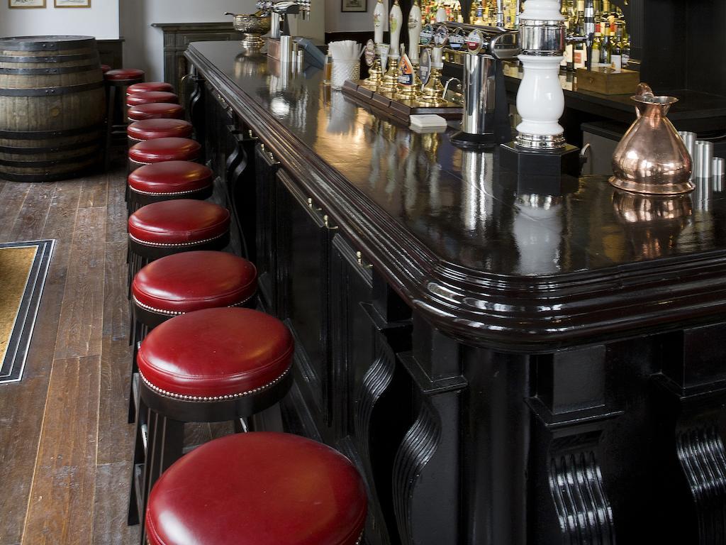 فندق Montcalm Brewery Townhouse لندن المظهر الخارجي الصورة The photo shows a bar setting with a long, polished dark wooden bar counter. Attached to the counter are several bar stools with red leather seats. Behind the bar, there are various bottles, suggesting a well-stocked bar. The flooring appears to be w