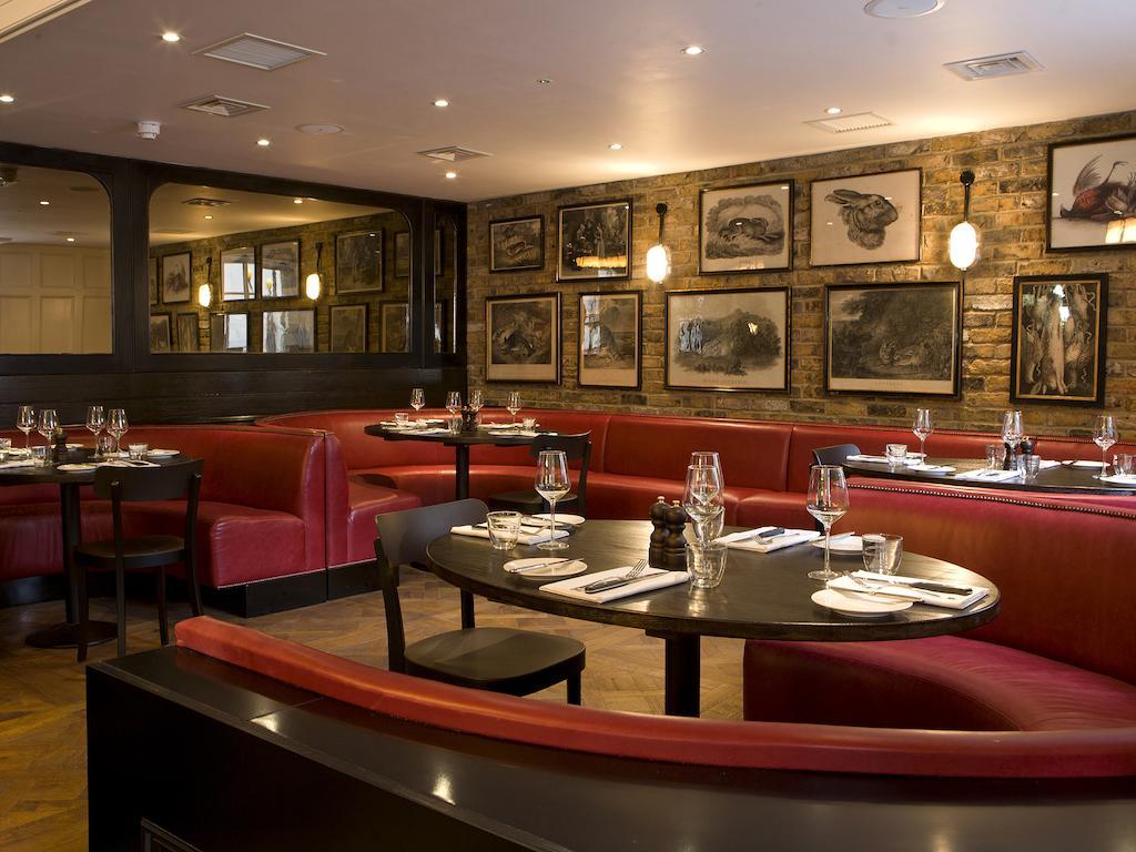فندق Montcalm Brewery Townhouse لندن المظهر الخارجي الصورة The photo depicts an elegant restaurant interior. There are curved red booths along the walls, creating a cozy seating arrangement. The tables are round and set with white plates and glassware, indicating a prepared dining area. The walls feature a n
