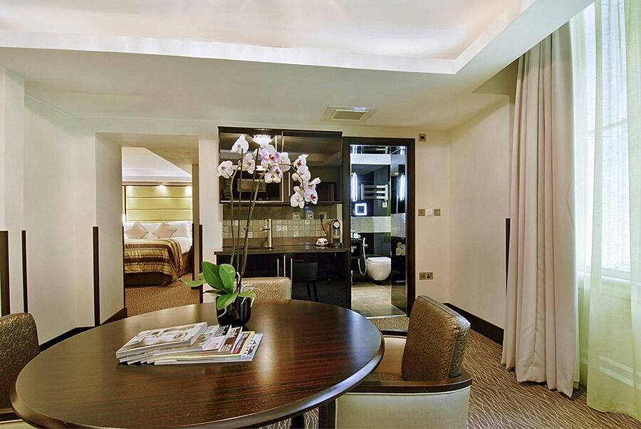 فندق Montcalm Brewery Townhouse لندن المظهر الخارجي الصورة The photo shows a stylish hotel room or suite. In the foreground, there is a round dining table with a few magazines on it. Surrounding the table are upholstered chairs. To the right, there are large windows with light-colored curtains, allowing natu