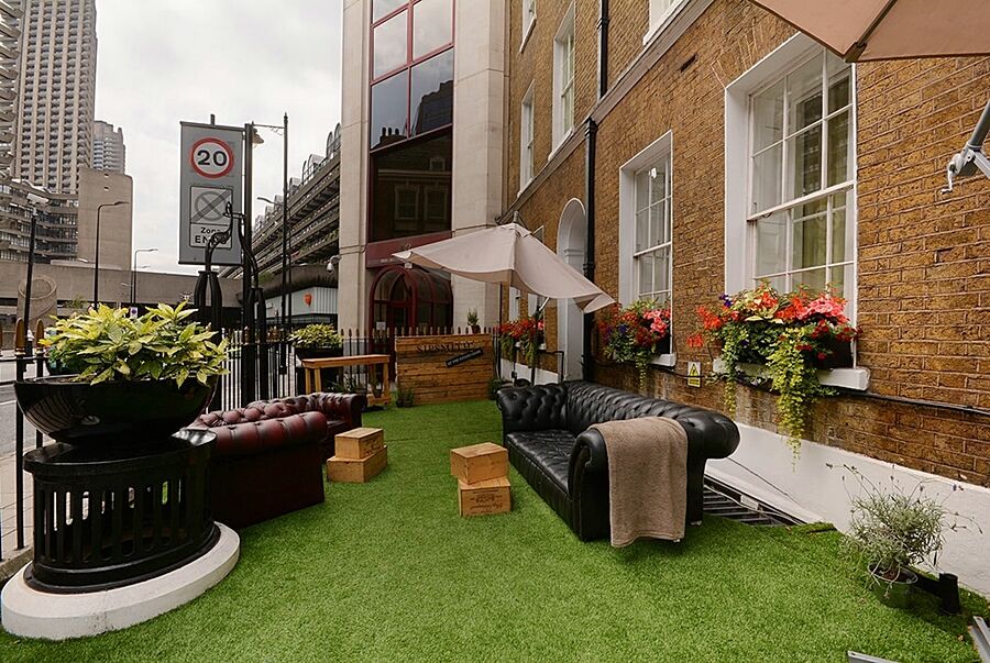 فندق Montcalm Brewery Townhouse لندن المظهر الخارجي الصورة The photo depicts a charming outdoor space, likely a patio or garden area. It features two comfortable-looking leather sofas, one of which is a deep burgundy color, placed on a green artificial lawn. There are wooden crates that could serve as side t