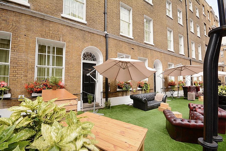 فندق Montcalm Brewery Townhouse لندن المظهر الخارجي الصورة The photo depicts an outdoor area of a building with a brick facade. There are several large windows visible, along with a wooden door. The setting features outdoor furniture, including couches in a dark leather finish and wooden tables. Large umbrel